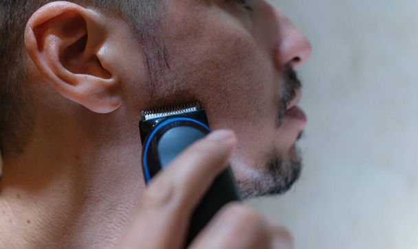 Close-up of the face of a young Caucasian man, using an electric razor to groom his sideburns. - Фото, изображение
