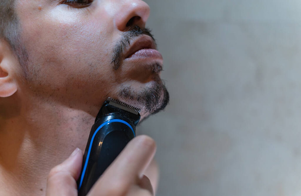 Zbliżenie na twarz młodego białego człowieka, używającego elektrycznej maszynki do golenia brody. - Zdjęcie, obraz