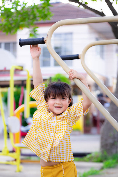 Retrato vertical. Menina bonita está se exercitando. As crianças asiáticas tentam estender a mão para pendurar em máquinas no playground. A criança ativa adora praticar esportes. Criança bonito com idade entre 5-6 anos. - Foto, Imagem