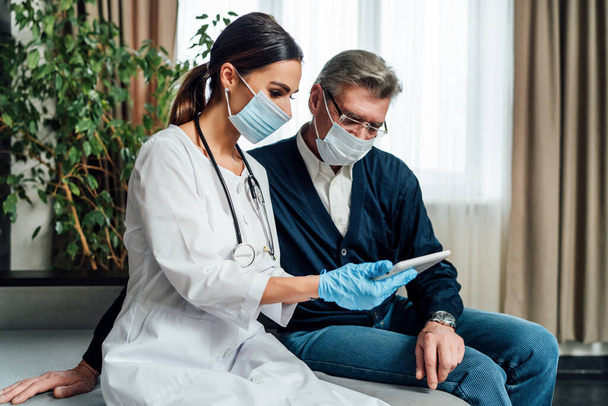 A nurse in a protective mask consults an older man at home, a senior man in a protective mask looks at the tablet. Virus protection, quarantine, home examinations. - Photo, Image