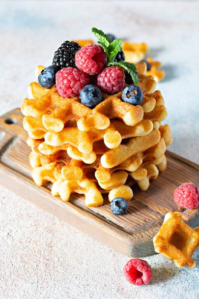 Fresh crispy Belgian waffles for breakfast with ripe berries (raspberries, blueberries, blackberries), mint and powdered sugar on a wooden board on a light background. - Photo, image