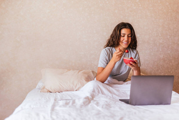Foto stock de menina feliz em pijama usando computador na cama e comer frutas. - Foto, Imagem