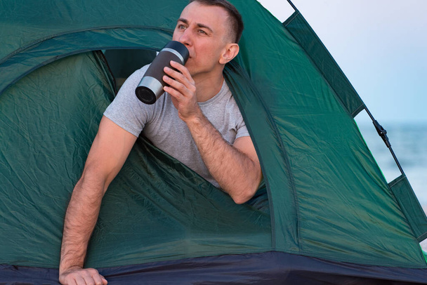 Un homme dans une tente boit une tasse de touriste. Randonnée matinale. Camping. - Photo, image