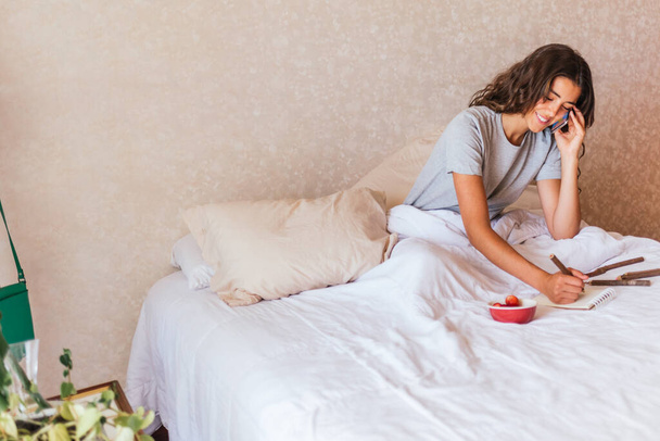 Photo de stock de fille heureuse en pyjama en utilisant son téléphone portable dans le lit et manger des fruits. - Photo, image
