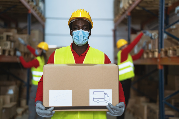 Homme noir travaillant dans des boîtes de livraison de chargement d'entrepôt tout en portant un masque chirurgical visage pendant une pandémie de virus corona - Concept logistique et de l'industrie - Photo, image