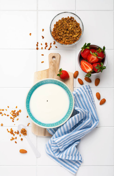 Kreikkalainen jogurtti valkoisessa kulhossa, jossa on ainesosia aamiaisen myslin ja tuoreiden mansikoiden valmistukseen valkoisella pöydällä. Yläkuva. - Valokuva, kuva