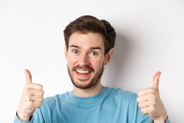 Close-up of cheerful man say yes, showing thumbs up in approval, praise good job, smiling approvingly, standing on white background - Photo, Image