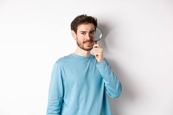 Smiling young man detective looking through magnifying glass, searching for promo offer, standing on white background - Photo, image