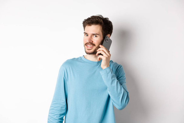 Technologiekonzept. Junges männliches Model spricht auf Handy, telefoniert mit jemandem auf Smartphone und lächelt vor weißem Hintergrund - Foto, Bild