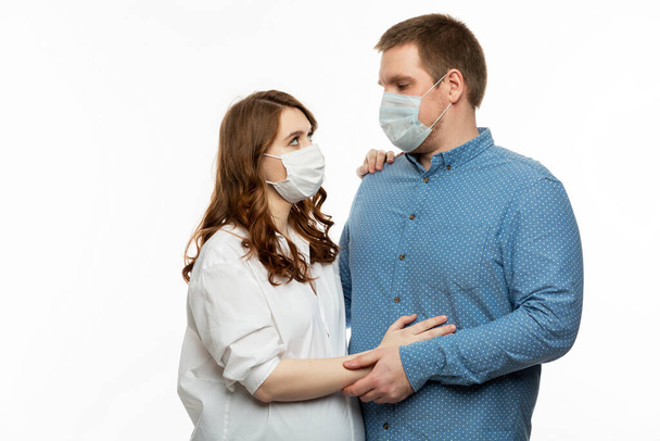 Young couple in medical masks. A pregnant woman with her husband are embracing. White background. - Φωτογραφία, εικόνα