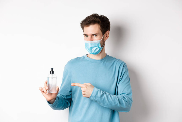 Covid-19, υγεία και καραντίνα έννοια. Καυκάσιος άνδρας με ιατρική μάσκα δείχνει το δάχτυλο σε μπουκάλι με απολυμαντικό χεριών, συνιστώντας αντισηπτικό, στέκεται σε λευκό φόντο - Φωτογραφία, εικόνα