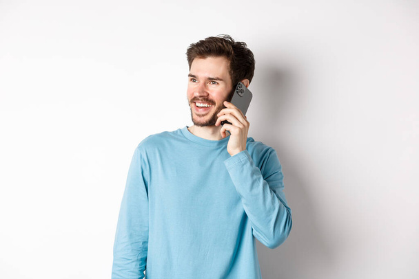 Technologiekonzept. Fröhlicher Mann, der das Telefonieren genießt, mit dem Smartphone spricht und lächelt, lässig im Hemd vor weißem Hintergrund steht - Foto, Bild