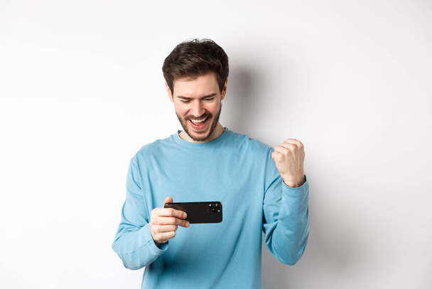 Щасливий молодий чоловік виграє в відеоіграх на смартфоні, дивлячись на мобільний екран і кажучи "так", роблячи кулачковий насос на святкуванні, досягає мети в Інтернеті, стоячи над білим тлом
 - Фото, зображення