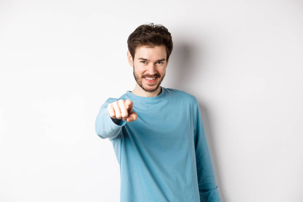 Image of confident young man smiling and pointing at camera, inviting and choosing you, beckon to join him, standing on white background - Photo, Image