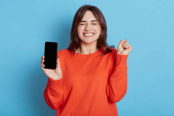 Overjoyed attractive woman keeps fist clenched, being happy to get good news, gets notification on cellphone, wears orange sweater, stands with closed eyes indoor against blue wall, feels amused. - Foto, imagen