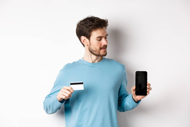 Мобільний банкінг. Красивий білий чоловік показує пластикову кредитну картку і дивиться на порожній мобільний екран, що стоїть на білому тлі
 - Фото, зображення