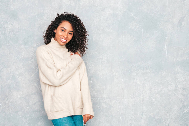 Όμορφη μαύρη γυναίκα με αφρο μπούκλες χτένισμα.Smiling μοντέλο σε λευκό μοντέρνο τζιν ρούχα. Σέξι ανέμελη γυναίκα που ποζάρει κοντά σε γκρίζο τοίχο στο στούντιο. Μαυρισμένος και χαρούμενος - Φωτογραφία, εικόνα