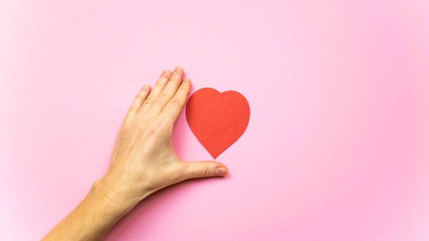 Herz zwischen den Fingern einer Frauenhand isoliert auf hellrosa Hintergrund. Valentinstag, Muttertag oder Hochzeit- und Liebeskonzept. - Foto, Bild