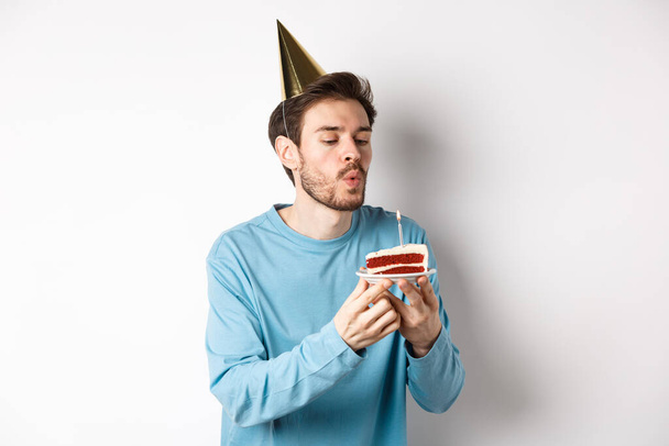 Oslava a svátky. Šťastný hlupák sfouknutí svíčky na dort a přání, stojící v klobouku nad bílým pozadím - Fotografie, Obrázek