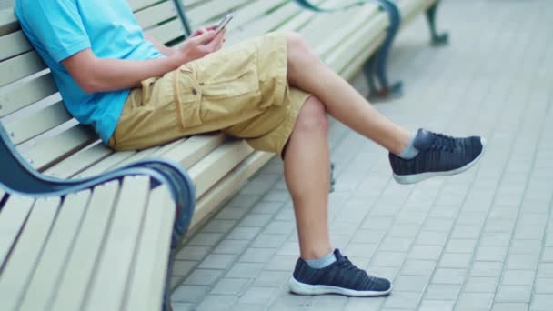 Szőrös lábak egy rövidnadrágos férfi ül egy padon egy mobiltelefon, kamera követés - Felvétel, videó