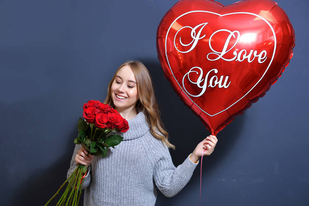 jeune fille blonde dans un pull gris tenant un bouquet de roses rouges et un ballon coeur. Joyeux concept de Saint-Valentin. jeune femme souriant sur fond gris - Photo, image