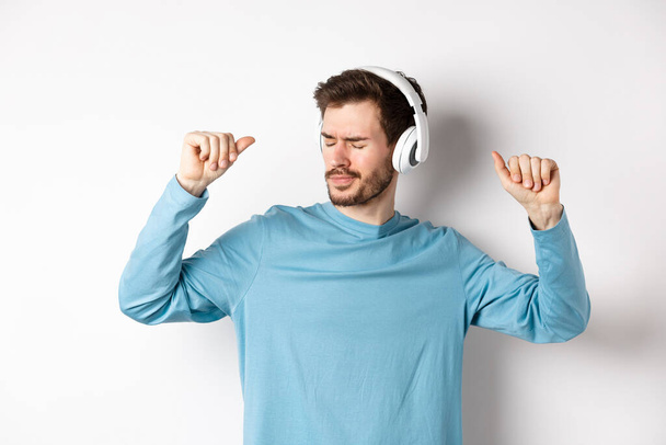 Szczęśliwy młodzieniec bawiący się w słuchawkach, tańczący podczas słuchania muzyki w bezprzewodowych słuchawkach, białe tło - Zdjęcie, obraz