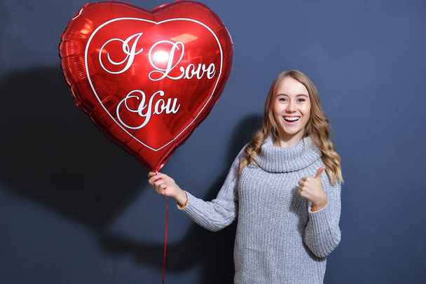 young blonde girl in a gray sweater holding a bouquet of red roses and a heart balloon. Happy valentine day concept. young woman smiling on gray background - Fotó, kép