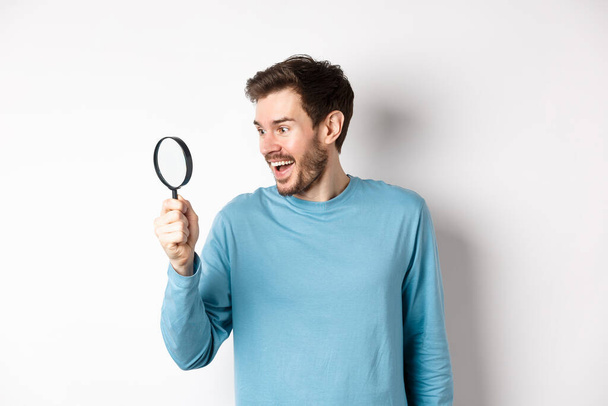 Handsome smiling man looking through magnifying glass with amazed face, found interesting promotion, standing on white background - Foto, afbeelding