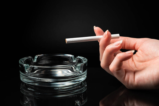 Eine Zigarette in der Hand einer Frau neben einem Aschenbecher auf schwarzem Hintergrund. - Foto, Bild