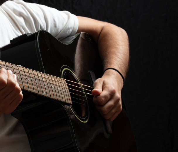 Ψυχαγωγία. Νεαρός που παίζει ακουστική κιθάρα, σκοτεινό φόντο. Guitarist σε casual λευκό πουκάμισο κατέχει ηλεκτρικό μαύρο όργανο στο στούντιο. - Φωτογραφία, εικόνα