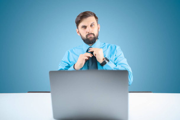 un hombre de negocios con una camisa azul ajusta el nudo de su corbata mientras está sentado en una computadora portátil, un hombre barbudo en la oficina - Foto, imagen