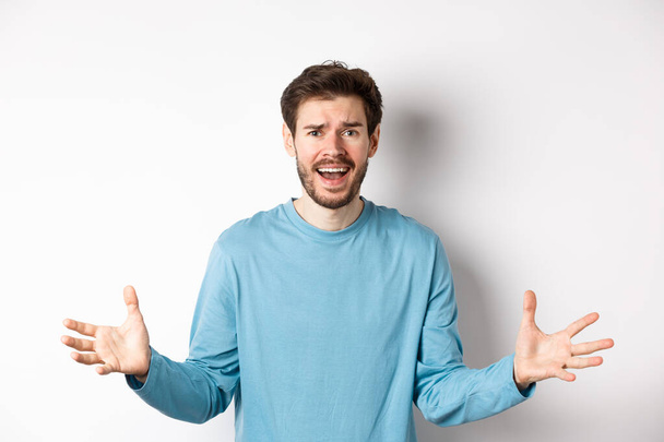 Image of frustrated and worried young man shaking hands, trying to explain something, standing anxious against white background - Фото, зображення