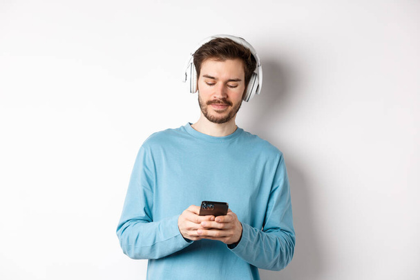 Modern young man in blue sweatshirt put on music on smartphone, listening songs in wireless headphones, white background - 写真・画像