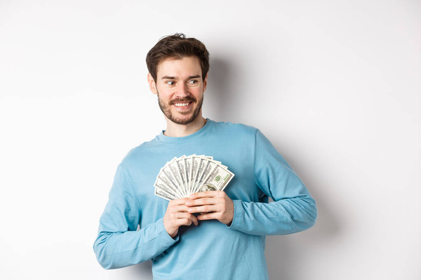 Chciwy uśmiechnięty mężczyzna pokazujący pieniądze i patrzący w prawo, myślący o zakupach, stojący na białym tle - Zdjęcie, obraz