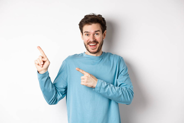 Homem caucasiano feliz em camisola azul casual, apontando os dedos no canto superior esquerdo, mostrando link ou logotipo no fundo branco, sorrindo para a câmera - Foto, Imagem