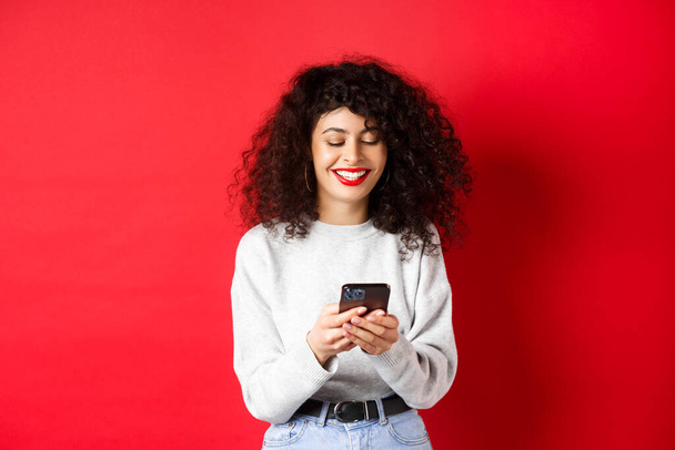Chica positiva feliz charlando por teléfono, leyendo el mensaje y sonriendo, utilizando la aplicación de medios sociales, de pie sobre fondo rojo - Foto, Imagen