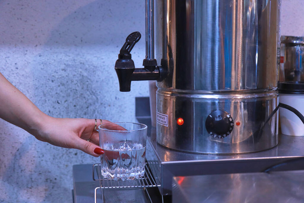 una mano de mujer presiona el grifo para verter agua caliente de la caldera en un vaso transparente sobre la mesa - Foto, Imagen