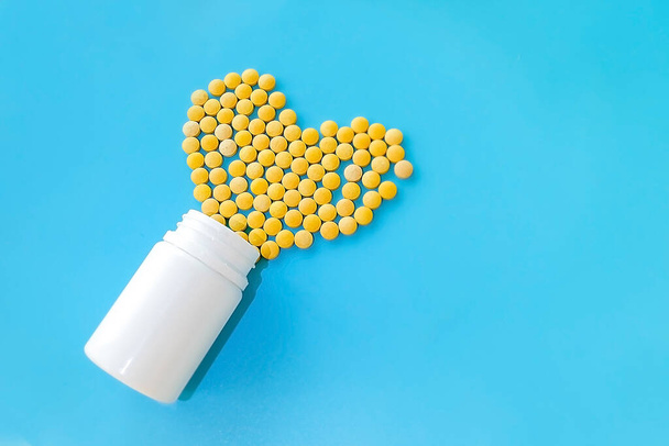many small yellow pills spilled out of a white jar on a blue background in the shape of a hear - Photo, Image