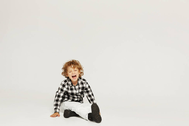 Crying curly little boy sits on the floor and keeping hand on his leg. A small kid broke his leg, isolated over gray background - Photo, image