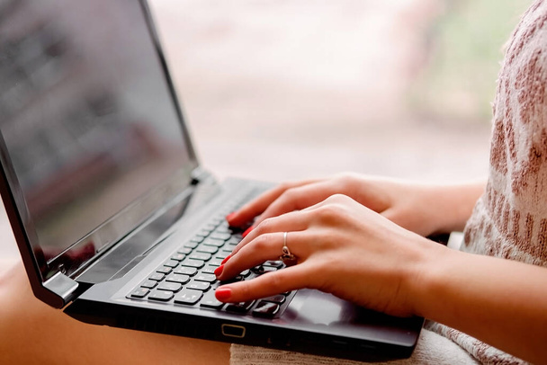menina em uma camisola de malha leve senta-se perto da janela e mantém um laptop. Mãos femininas imprimir em um notebook, trabalho remoto em hom - Foto, Imagem