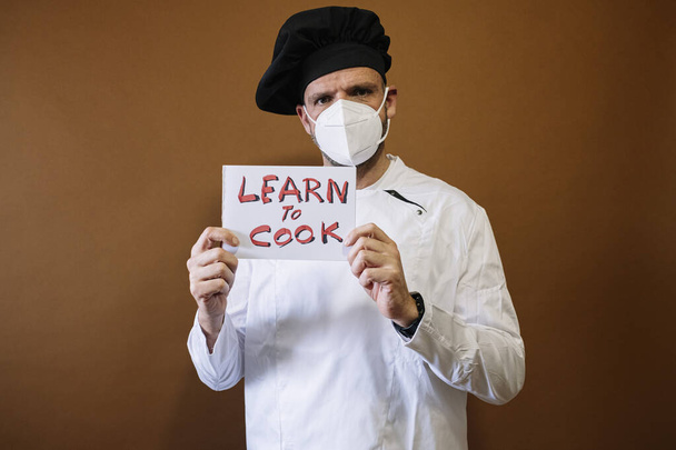 Шеф-повар держит табличку с надписью "учиться и готовить", носит маску на лице. - Фото, изображение