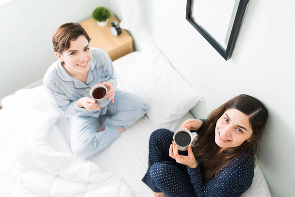 Two young women friends with pajamas sitting in bed and holding coffee cups. Smiling best friends enjoying a sunny morning in the bedroom - Photo, Image