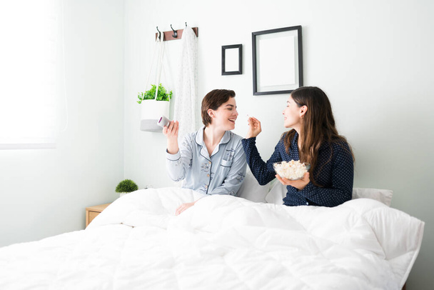 Beautiful smiling latin woman feeding popcorn to her pretty young girlfriend while sitting in bed. Two women in a relationship watching a movie in pajamas - Photo, Image