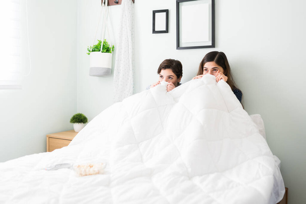 Bang beste vriendinnen kijken naar een horrorfilm in bed en verstoppen zich onder de dekens. Twee vrouwen vrienden hebben plezier met een eng tv-serie tijdens een logeerpartijtje - Foto, afbeelding