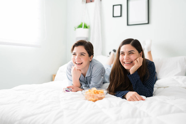 Happy female best friends laughing while watching a comedy movie in bed. Pretty young women eating a bowl of chips in pajamas during a fun slumber party - Photo, Image