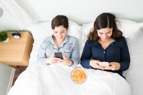 Beautiful young women friends laughing and lying in bed during a sleepover. Top view of two best friends texting and scrolling through social media in their smartphones - Foto, afbeelding