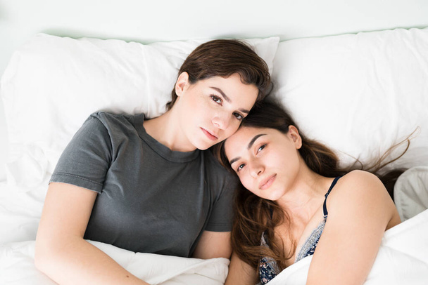 Porträt eines heiteren schwulen lesbischen Paares, das morgens in seinem Schlafzimmer aufwacht. Schöne lateinische und kaukasische Frauen, die sich im Bett umarmen  - Foto, Bild