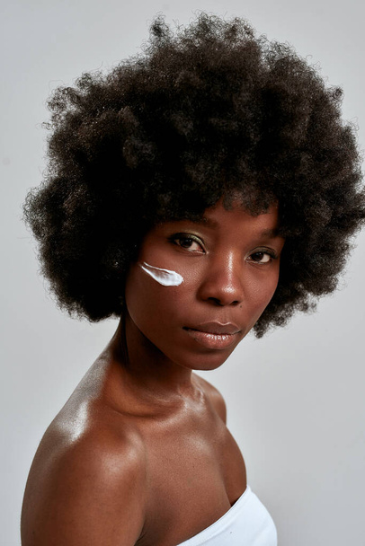Beauty portrait of attractive young naked african american woman with afro hair looking at camera while posing with cream applied on her face isolated over gray background - Valokuva, kuva