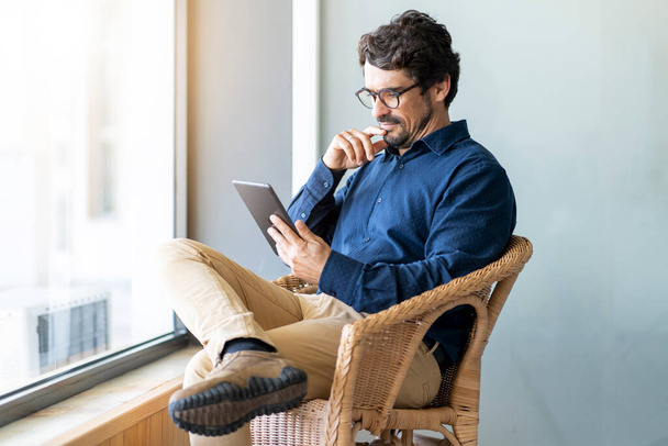 Hombre de negocios casual con gafas sentado mirando su tableta. Retrato masculino exitoso con expresión de cara pensante trabajando desde casa junto a la ventana - Foto, imagen