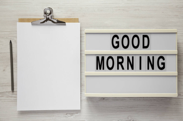 'Good morning' on a lightbox, clipboard with blank sheet of paper on a white wooden background, top view. Flat lay, overhead, from above.  - Photo, Image